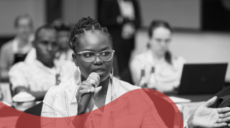 PPhoto of a black woman holding a microphone while speaking in a room with other people. 