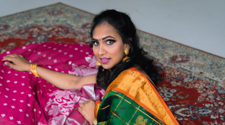 A woman with dark hair, in colorful attire poses sitting on the floor and looking directly at the camera. She's sitting on a rug on the floor and her eyes are highlighted with eyeliner. 